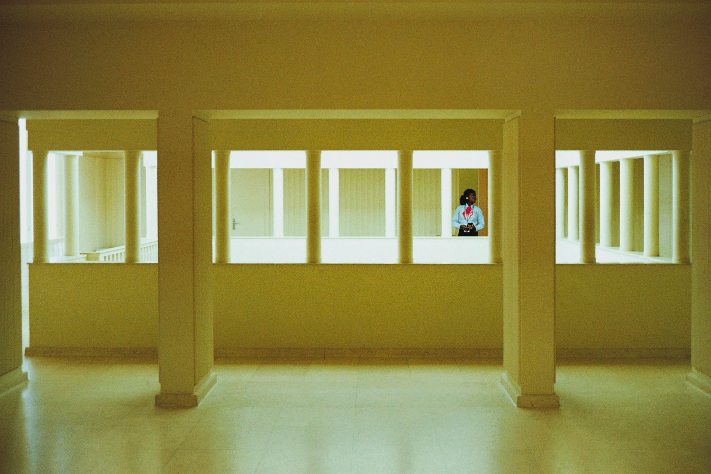 Warm yellow Art Deco interior of Casa de Serralves, where square pillars frame a colonnaded gallery and a woman in a pale blue shirt stands alone at the far side.