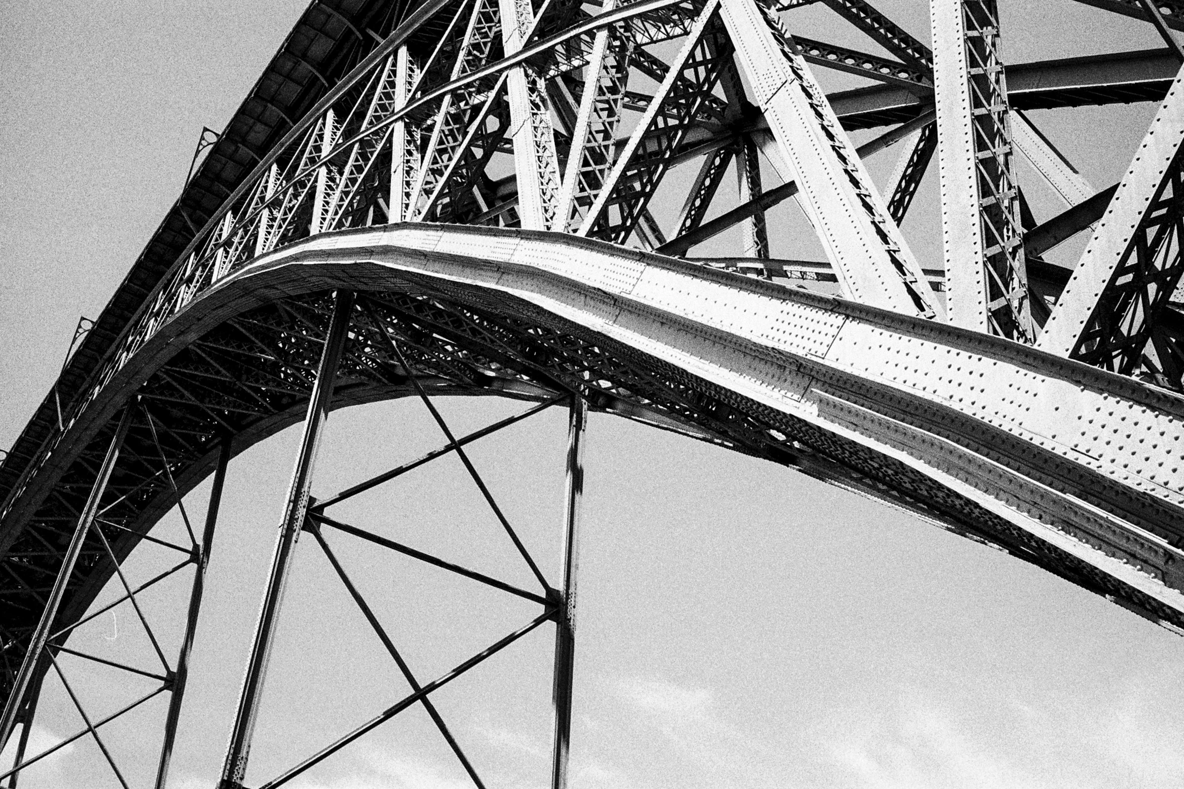 Black-and-white view from beneath the Ponte Luís I, its riveted iron arch and lattice girders crossing a pale sky.