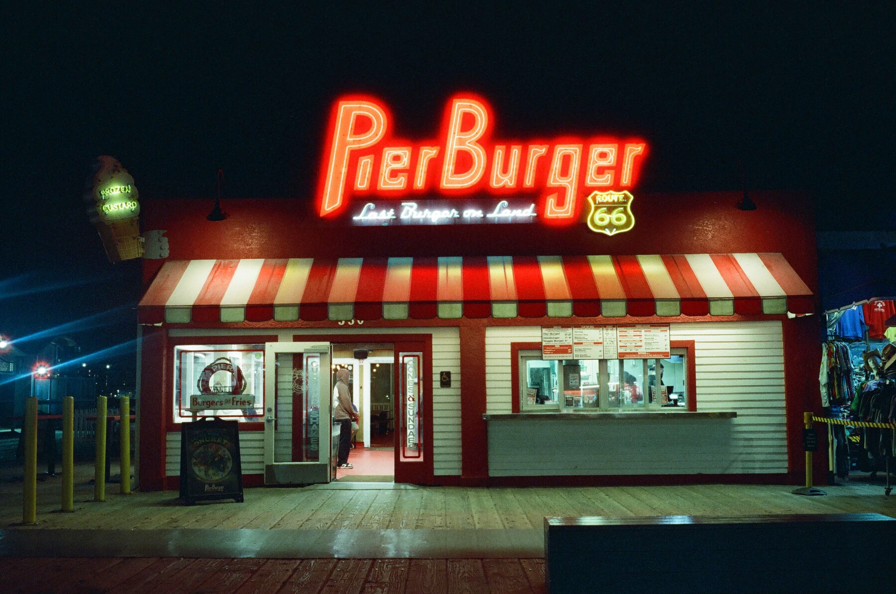 Pier Burger at night on the Santa Monica Pier: a red neon sign reading “Last Burger on Land” and a Route 66 shield above a red-and-white striped awning.