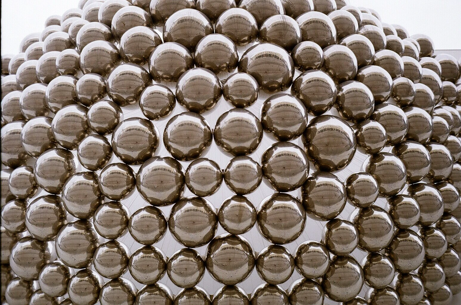 A wall of hundreds of mirrored steel spheres of different sizes, each reflecting the buildings and pale sky around it.
