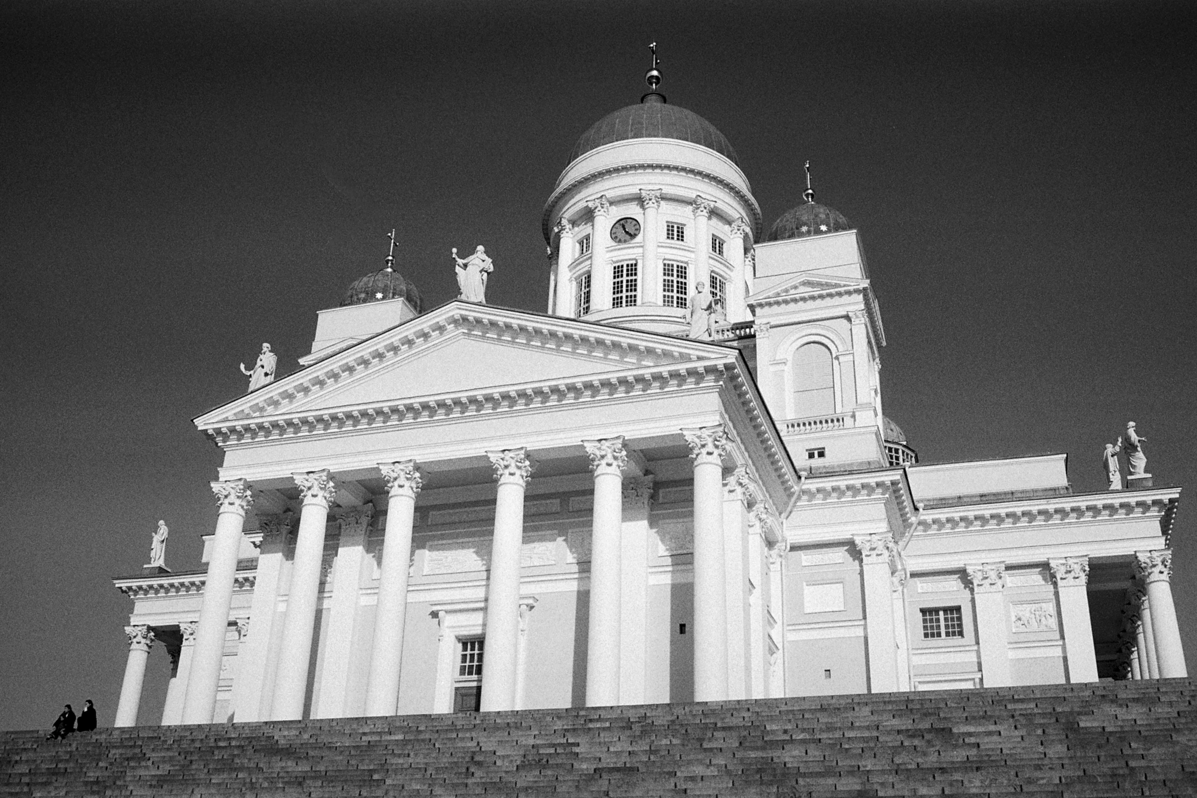 Black-and-white photo of Helsinki Cathedral's white columns, statues, and domes seen from the foot of its steps, with two small figures seated at the left.