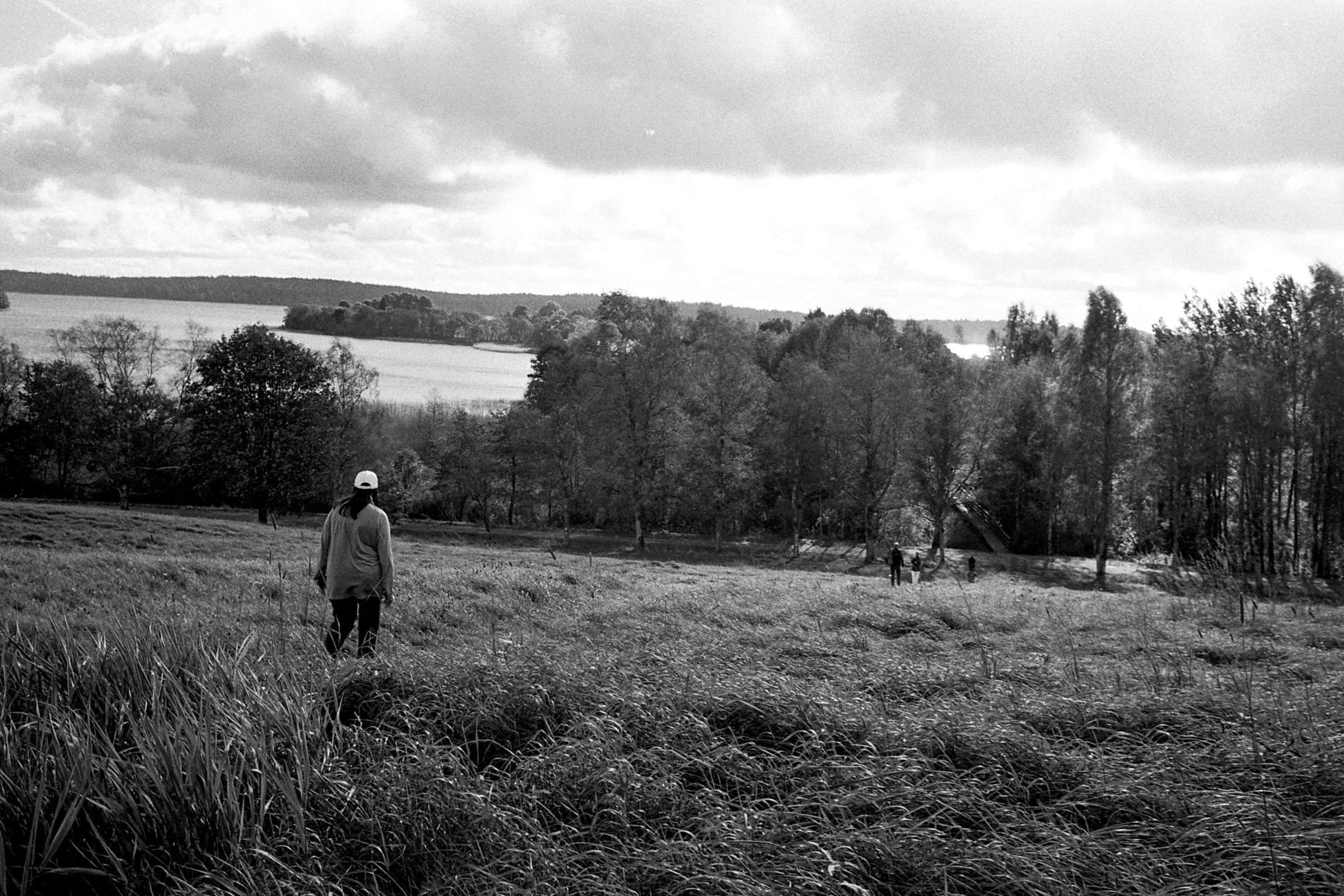 Black-and-white photo of a person in a cap walking down a grassy meadow toward trees and a wide lake, with a few distant figures below and clouds above.