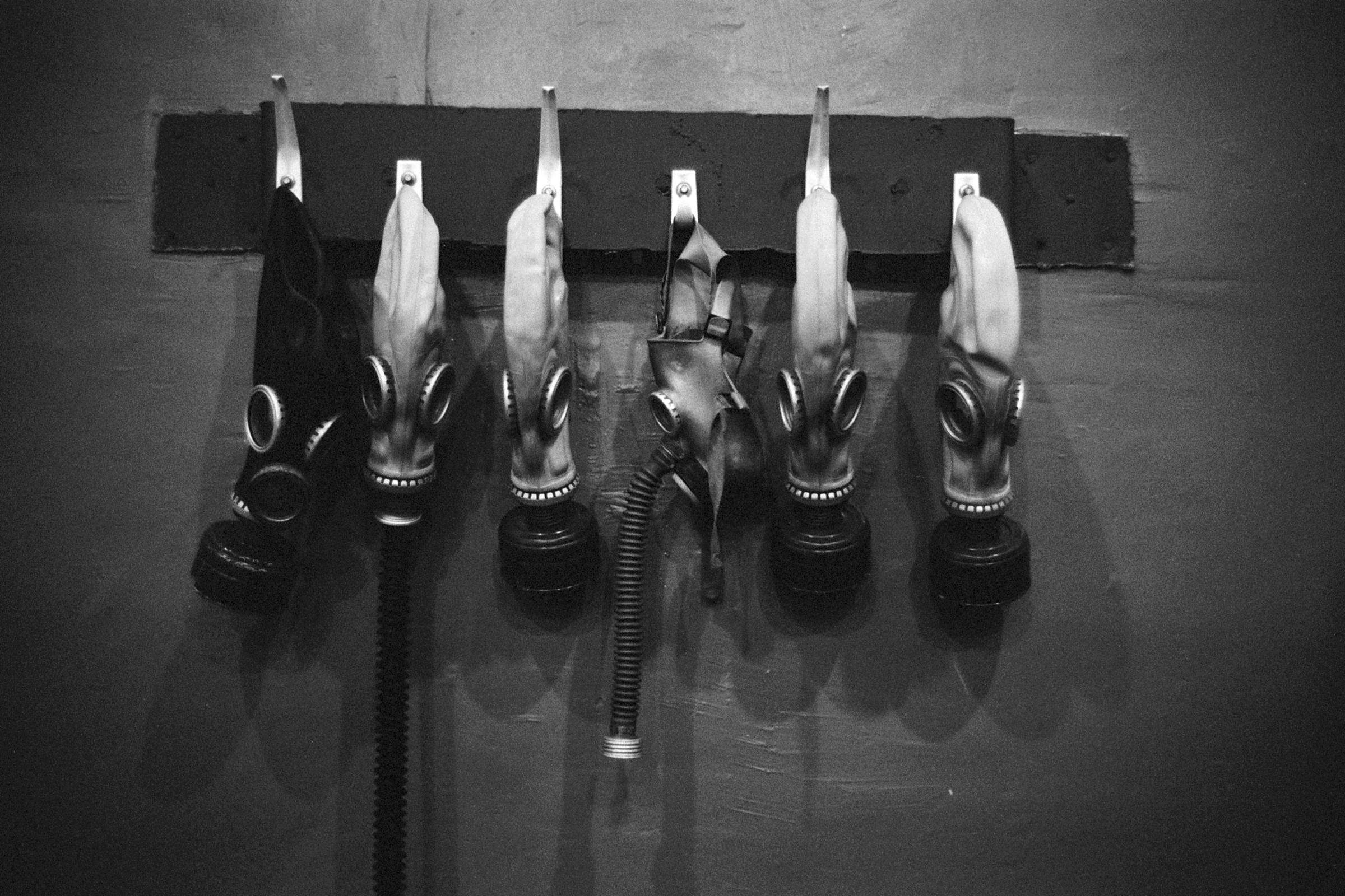 Black-and-white photo of six Soviet gas masks hanging from hooks on a wall, one crumpled with its breathing hose hanging down.
